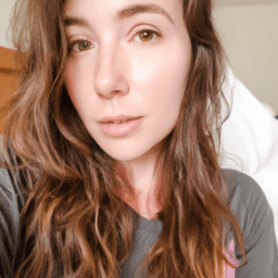 A photograph of an attractive woman, freshly awakened, capturing a home-made style selfie. She looks into the camera with sleepy yet content eyes. Background is a cozy bedroom with white linens. Soft, dawn light filters through sheer curtains, casting a golden glow on her face.